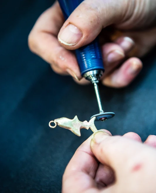 A jeweller using a rotary tool to refine and polish a handmade fish-shaped metal pendant, focusing on the intricate details. A jeweller using a rotary tool to refine and polish a handmade fish-shaped metal pendant, focusing on the intricate details.
